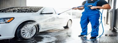 A man in blue overalls washing a white car with a high-pressure washer. Car wash business in UAE.